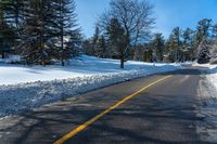 a snowy street that is next to a forest and snow covered hill, with a yellow line in the middle