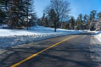 a snowy street that is next to a forest and snow covered hill, with a yellow line in the middle