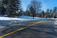 a snowy street that is next to a forest and snow covered hill, with a yellow line in the middle