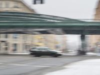 the car is driving on the street in the snow past a green bridge and buildings