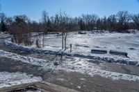 a park with snow around a wooden bench and trees in the distance, on a sunny day