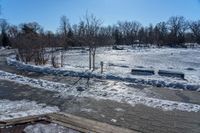 a park with snow around a wooden bench and trees in the distance, on a sunny day