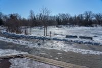 a park with snow around a wooden bench and trees in the distance, on a sunny day