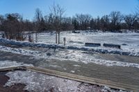 a park with snow around a wooden bench and trees in the distance, on a sunny day