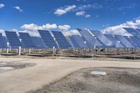 rows of solar panels line a dirt road at a construction site, under a blue sky and clouds