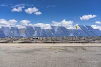 rows of solar panels sit on a paved area against a blue sky with white clouds