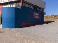 the red and blue building is set in dirt area and it has street signs on top of it