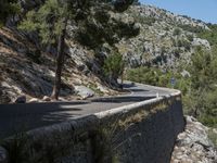 a road with a long curve through the mountains, with trees and a rock wall beside it