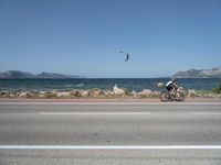 man riding bike along side the water with kite in sky above him on shore line