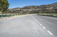 two lane paved road and mountain range in distance with trees on either side and green fence