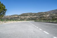 two lane paved road and mountain range in distance with trees on either side and green fence