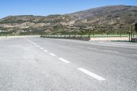 two lane paved road and mountain range in distance with trees on either side and green fence