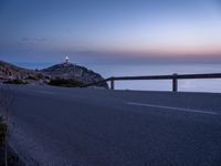 the street is near a mountain and looks out at the sea at dusk with a lighthouse on the horizon