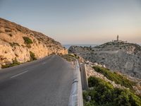 a winding road on a cliff overlooking a sea and a lighthouse tower at sunset, a view from behind