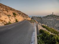 a winding road on a cliff overlooking a sea and a lighthouse tower at sunset, a view from behind