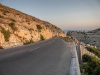 a winding road on a cliff overlooking a sea and a lighthouse tower at sunset, a view from behind