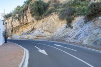 the paved street in the city is next to a cliff with bushes on top of it