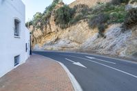 the paved street in the city is next to a cliff with bushes on top of it