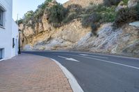 the paved street in the city is next to a cliff with bushes on top of it