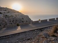 a small car is driving down a road beside a stone cliff by the water during sunset