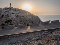 a small car is driving down a road beside a stone cliff by the water during sunset