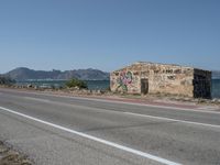 a graffiti covered building is by the edge of a beach road by the ocean in a coastal area