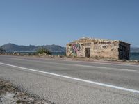 a graffiti covered building is by the edge of a beach road by the ocean in a coastal area