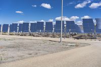 several rows of large solar panels against a bright blue sky with clouds and water behind them