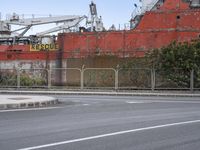 a rusty ship sits in the distance next to a fence on a street corner where a red bus is parked