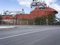 a rusty ship sits in the distance next to a fence on a street corner where a red bus is parked