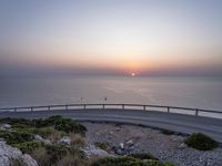 a curved road going to the ocean on a clear day with a bright sun rising over the horizon