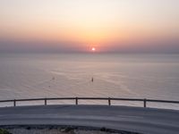 a curved road going to the ocean on a clear day with a bright sun rising over the horizon