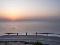 a curved road going to the ocean on a clear day with a bright sun rising over the horizon