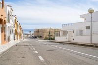 a street with some white buildings on one side and blue ocean in the distance and sky and buildings to the left