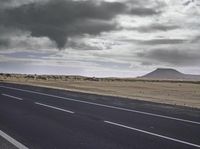 the asphalt road leading towards the desert mountain under a cloudy sky with white clouds above