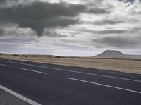 the asphalt road leading towards the desert mountain under a cloudy sky with white clouds above