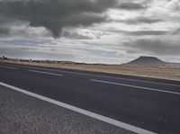 the asphalt road leading towards the desert mountain under a cloudy sky with white clouds above