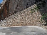 a man riding a motorcycle down a dirt road near a large rock wall and stone staircase