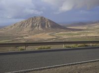 a road with mountain and valley in the background, surrounded by grass, shrubs and dirt