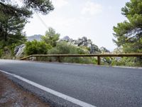 an empty asphalt road running along side some trees and rocks along a fence line and mountains