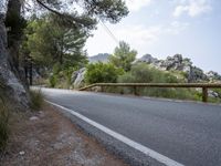 an empty asphalt road running along side some trees and rocks along a fence line and mountains