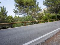 an empty asphalt road running along side some trees and rocks along a fence line and mountains