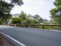 an empty asphalt road running along side some trees and rocks along a fence line and mountains