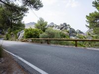 an empty asphalt road running along side some trees and rocks along a fence line and mountains