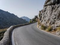 an empty asphalt road winds through a rocky area with mountains in the background and blue sky overhead