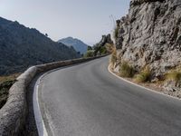 an empty asphalt road winds through a rocky area with mountains in the background and blue sky overhead
