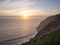 a person sitting on a hill overlooking the ocean and ocean waves at sunset time looking out over the ocean