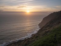 a person sitting on a hill overlooking the ocean and ocean waves at sunset time looking out over the ocean