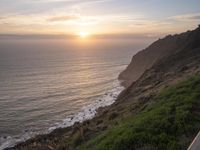 a person sitting on a hill overlooking the ocean and ocean waves at sunset time looking out over the ocean