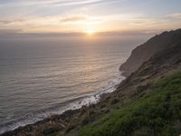 a person sitting on a hill overlooking the ocean and ocean waves at sunset time looking out over the ocean